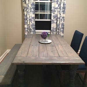 May include: A farmhouse style dining table with a gray stained wood top and black painted legs. The table has a bench on one side and two chairs on the other side. The table is set with a vase of purple flowers.