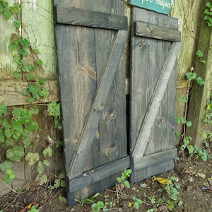 May include: Two dark gray wooden shutters with a diagonal brace and a black painted finish. The shutters are leaning against a green wooden fence with a sign that reads "Unique Primitives".