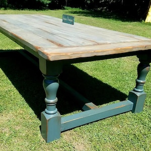 May include: A large rectangular wooden table with a weathered light gray top and a dark blue-green base featuring four turned legs and a central support beam. A small rectangular sign with the text Unique & Boutique sits on the table surface.