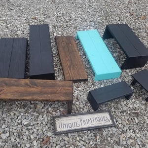 May include: A collection of wooden shelves in various sizes and colors, including black, brown, and turquoise. The shelves are made of rustic wood and have a simple, farmhouse style. The shelves are displayed on a gravel surface with a sign that reads "Unique Primitives".
