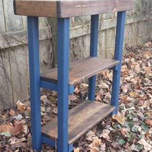 May include: A wooden shelf unit with three shelves, painted in a dark blue color. The shelves are made of natural wood with a rustic finish. The unit is standing on a bed of fallen leaves.