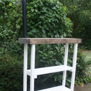 May include: White wooden shelf unit with two shelves and a dark brown stained top. The shelf unit is standing on a concrete surface in front of a green bush.