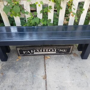 May include: A dark navy blue wooden bench with a rectangular seat and four sturdy legs. The bench is positioned in front of a white picket fence with green vines. A sign below the bench reads "UNIQUE PRIMITIQUES FARMHOUSE RUSTIC FURNITURE EST. 2010."
