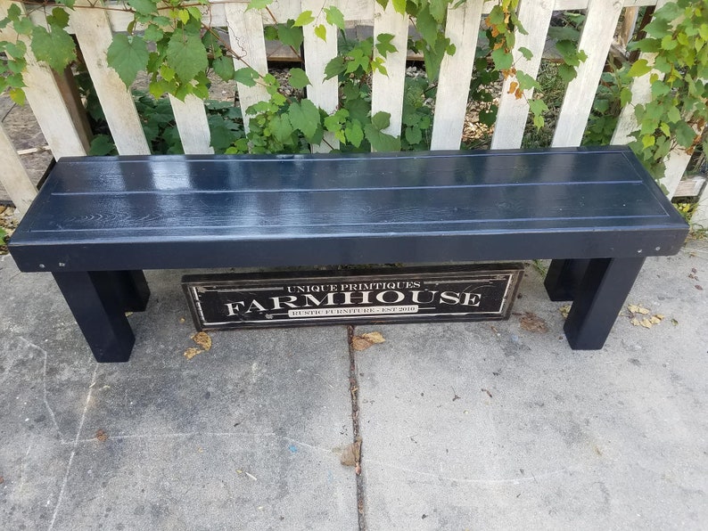 May include: A dark navy blue wooden bench with a rectangular seat and four sturdy legs. The bench is positioned in front of a white picket fence with green vines. A sign below the bench reads "UNIQUE PRIMITIQUES FARMHOUSE RUSTIC FURNITURE EST. 2010."