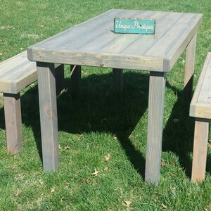 May include: A rustic wooden table and two benches set on a grassy lawn. The table is gray with a weathered finish and has a sign that reads "Unique  Prestiques". The benches are also gray and weathered.