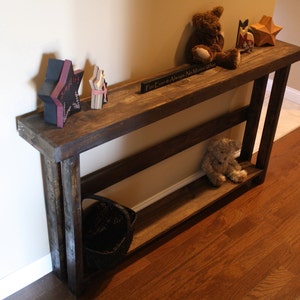 May include: A rustic wooden console table with two shelves. The table is stained a dark brown and has a distressed finish. The table is decorated with a variety of items, including a stuffed teddy bear, a star-shaped ornament, and a sign that reads "For Ever & Always No Matter What".
