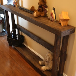 May include: A dark brown wooden console table with two shelves. The table has a rustic farmhouse style and is decorated with a teddy bear, a star, and other decorative items. The table is sitting on a hardwood floor.