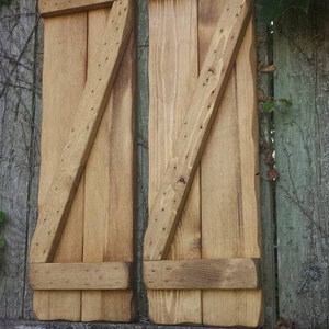May include: Two wooden shutters with a diagonal design, painted in a light brown color. The shutters are attached to a weathered wooden fence.