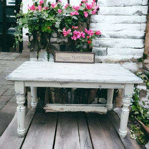 May include: A distressed white wooden table with turned legs. The table is placed on a wooden deck. A sign that reads "UNIQUE PRIMITIQUES" is on the table. Pink flowers and greenery are in a planter behind the table.