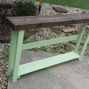 May include: A wooden console table with a dark brown top and light green legs. The table has a lower shelf and is designed for placement against a wall. The table is made of wood and is suitable for indoor or outdoor use.