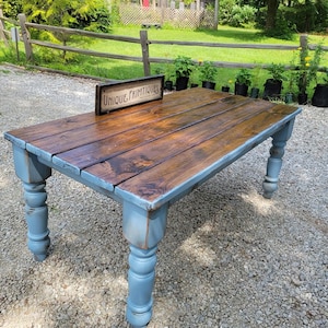 May include: A rustic wooden table with a stained brown tabletop and light blue painted legs. The table has a rectangular shape and is set outdoors on a gravel surface. A sign that reads "UNIQUE PRIMTIQUES" is on the table.