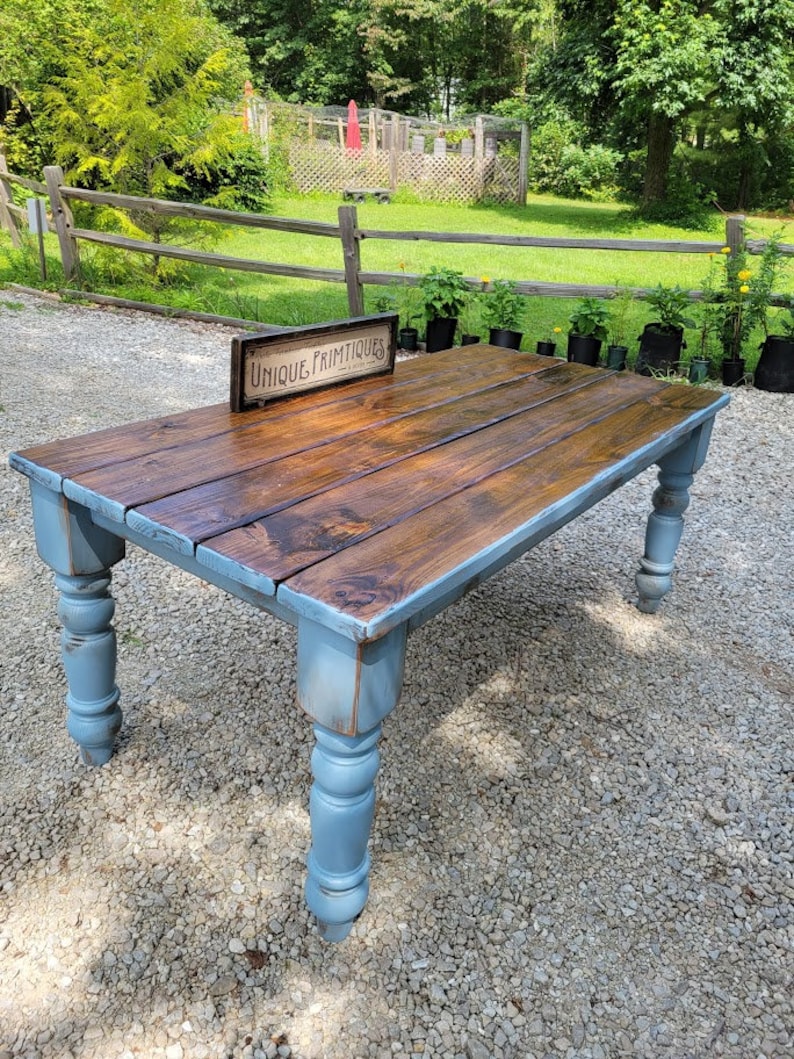 May include: A rustic wooden table with a stained brown tabletop and light blue painted legs. The table has a rectangular shape and is set outdoors on a gravel surface. A sign that reads "UNIQUE PRIMTIQUES" is on the table.