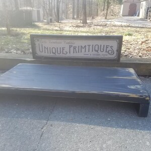 May include: A black wooden shelf with a distressed finish. The shelf is sitting on a gray concrete surface. A sign above the shelf reads "Rustic Farmhouse Treasures Unique Primitives & Decor".