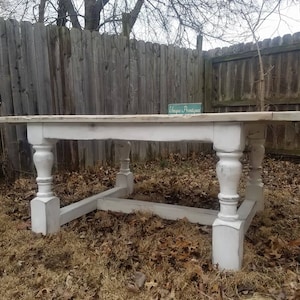 May include: A rectangular wooden table featuring a natural wood-toned top and a distressed white painted base. The table stands on four thick, turned legs connected by a sturdy lower frame. A small green sign with the text Unique Boutiques is visible.