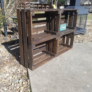 May include: A set of four wooden crates stacked to create a shelf unit. The crates are stained a dark brown color and have a rustic appearance. The top crate has a small sign that reads "Pristine".