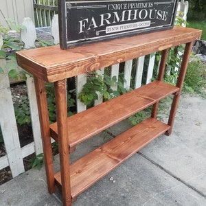 May include: A rustic wooden console table with two shelves. The table is stained a dark brown color and has a farmhouse style. The table is topped with a black sign that reads "Unique Primitives Farmhouse Rustic Furniture Est 2010".