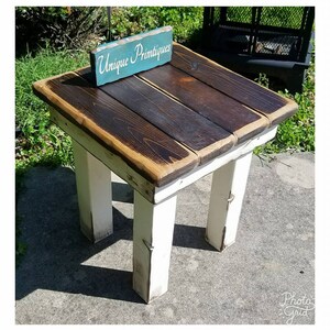 May include: A small wooden side table with four white painted legs and a dark brown stained plank top. A small teal rectangular sign with the white text Unique Primtiques is resting on the surface of the table. The table has a square top.
