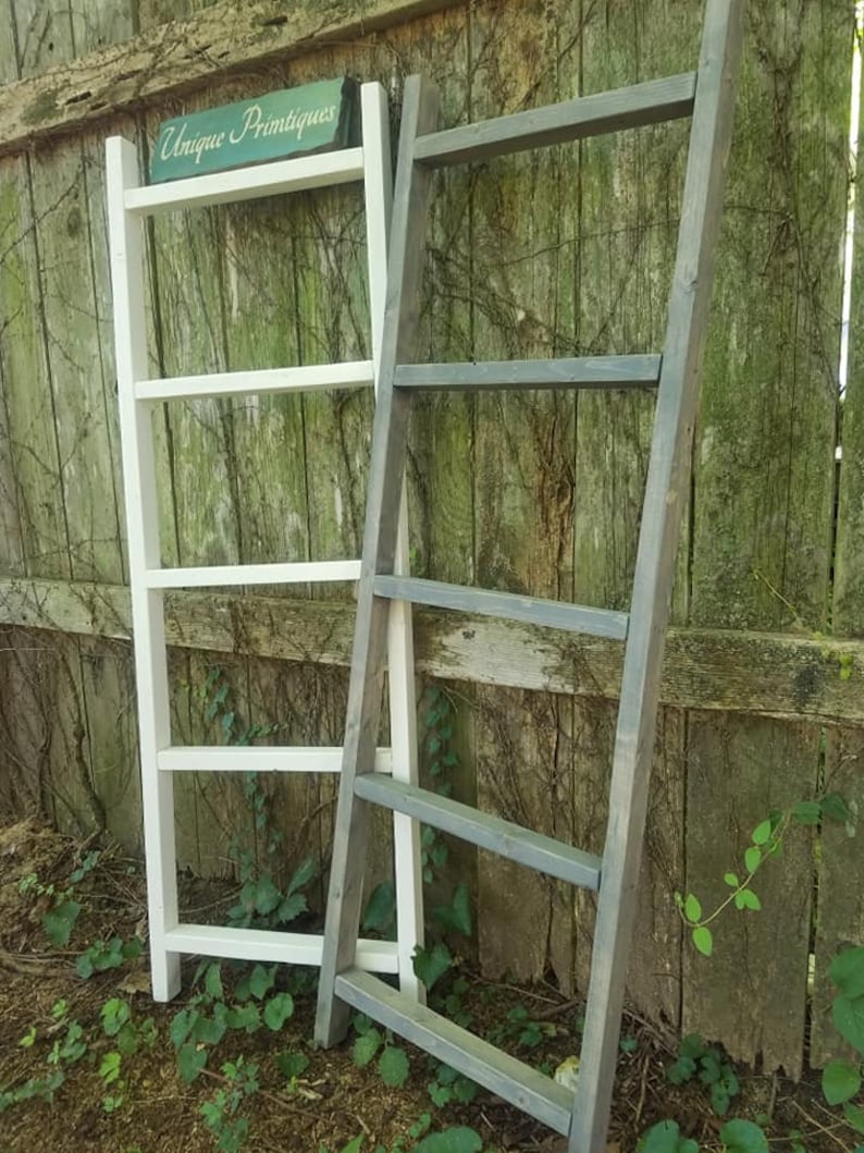 May include: Two wooden ladder shelves, one white and one gray, leaning against a wooden fence. The white ladder shelf has a sign that reads "Unique Printiques".