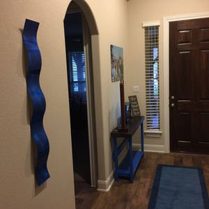 May include: A blue metal wall sculpture with a wavy design hangs on a beige wall in a hallway. A wooden table with a blue base sits in the hallway. A blue rug is on the floor. The text "Hope, Faith, Love" is on a sign on the table.