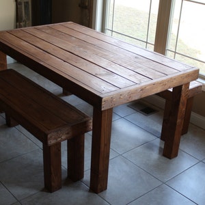 May include: A rustic wooden table and two benches set in a room with tiled floors. The table and benches are stained a dark brown color.