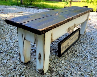 May include: A wooden bench featuring a dark stained top and a distressed white painted base with four sturdy legs. Positioned underneath the bench is a rectangular black and white sign with the text UNIQUE PRIMTIQUES printed on the front surface.