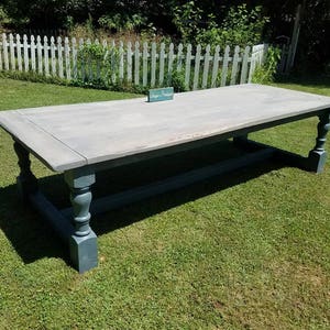 May include: A large rectangular wooden dining table featuring a light gray, weathered-look tabletop and a dark blue-gray base with four turned, pedestal-style legs connected by a central horizontal support beam. A small teal sign reads Unique Boutique.