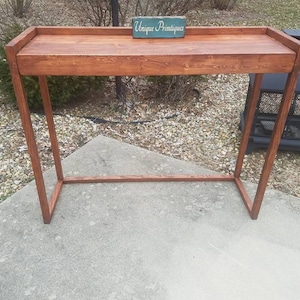 May include: A wooden console table with a brown stain finish. The table has a rectangular top and a simple, open frame. A small wooden sign with the text "Unique  Primitives" is placed on the table top.