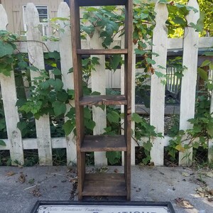 May include: A dark brown wooden shelf unit with four shelves. The shelf unit is standing in front of a white picket fence with green vines growing on it. A sign with the text "Rustic Farmhouse Furniture Unique Primitives & D&eacute;cor" is in front of the shelf unit.