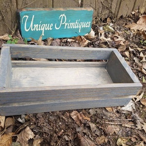 May include: A rectangular, gray-stained wooden crate with a slatted design. The crate has a light-colored interior and is set against a backdrop of fallen leaves and a wooden sign that reads "Unique Primitives" in white lettering on a teal background.