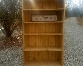 May include: A tall, unfinished wooden bookshelf with four shelves. The bookshelf has a rectangular shape and is made of light-colored wood. A sign that reads "UNIQUE PRIMTIQUES" is mounted on the middle shelf. The bookshelf is outdoors on a gravel surface.