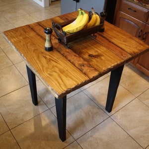 Puede incluir: Una mesa cuadrada de madera con patas negras. La superficie de la mesa est&aacute; hecha de madera recuperada y tiene un acabado natural. Un taz&oacute;n de pl&aacute;tanos se encuentra sobre la mesa.