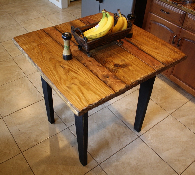Puede incluir: Una mesa cuadrada de madera con patas negras. La superficie de la mesa est&aacute; hecha de madera recuperada y tiene un acabado natural. Un taz&oacute;n de pl&aacute;tanos se encuentra sobre la mesa.