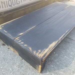 May include: A black wooden shelf with distressed edges and a light brown stain. The shelf is made of two planks of wood and has a rustic look.