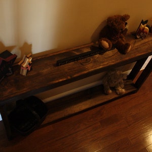 May include: A rustic wooden console table with a dark brown finish. The table has two shelves and is decorated with a variety of items, including a stuffed bear, a wooden star, and a sign that reads "Part of a Whole No Matter What".