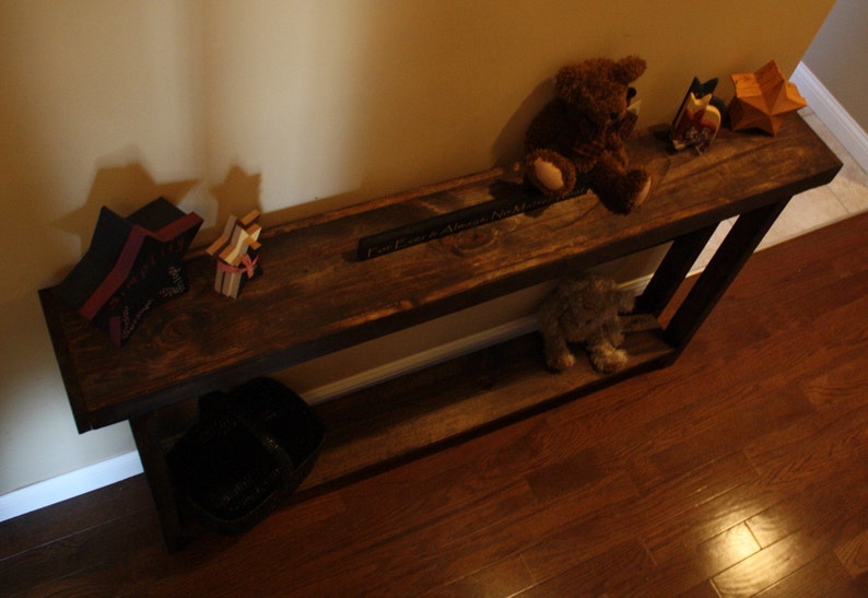 May include: A rustic wooden console table with a dark brown finish. The table has two shelves and is decorated with a variety of items, including a stuffed bear, a wooden star, and a sign that reads "Part of a Whole No Matter What".