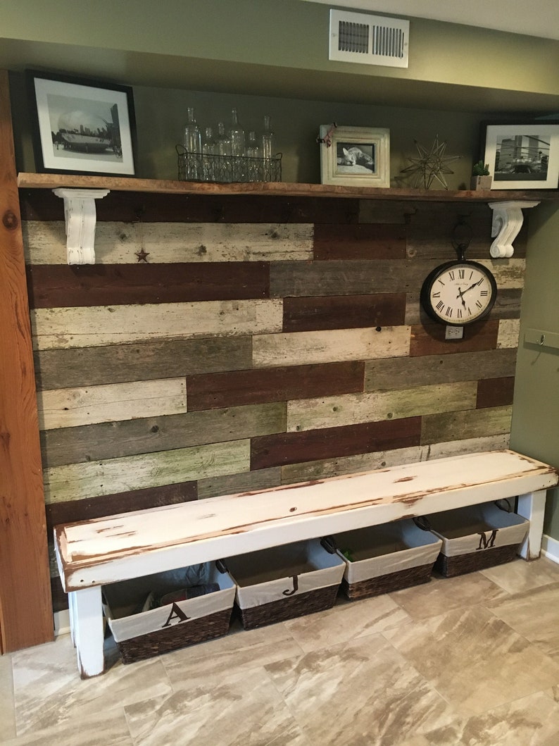 May include: A rustic, distressed white bench with three woven baskets, each labeled with a letter. The bench is set against a wall of reclaimed wood planks in shades of brown, white, and green. A shelf above displays framed photos and decorative items.