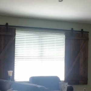 May include: A window with white blinds and two brown wooden barn door style shutters. The shutters are closed and have a black metal track system.