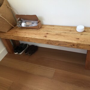 May include: A rustic wooden bench with a natural finish, featuring visible wood grain and sturdy legs. A woven basket, a cardboard box, and a pair of shoes are placed near the bench. A white, round object sits on the bench.