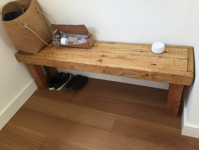May include: A rustic wooden bench with a natural finish, featuring visible wood grain and sturdy legs. A woven basket, a cardboard box, and a pair of shoes are placed near the bench. A white, round object sits on the bench.