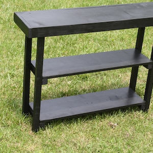 May include: A black wooden shelf unit with three shelves. The shelf unit is standing on a green grassy surface.