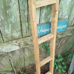 May include: A wooden ladder shelf with four rungs. The ladder is leaning against a wooden fence with green vines growing on it. A blue sign with the text "Unique Antiques" is visible in the background.
