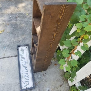 May include: A dark brown wooden shelf unit with four shelves. The shelf unit is standing on a gray concrete surface. A sign that reads "Unique Primitives - Home Decor" is lying on the ground in front of the shelf unit.