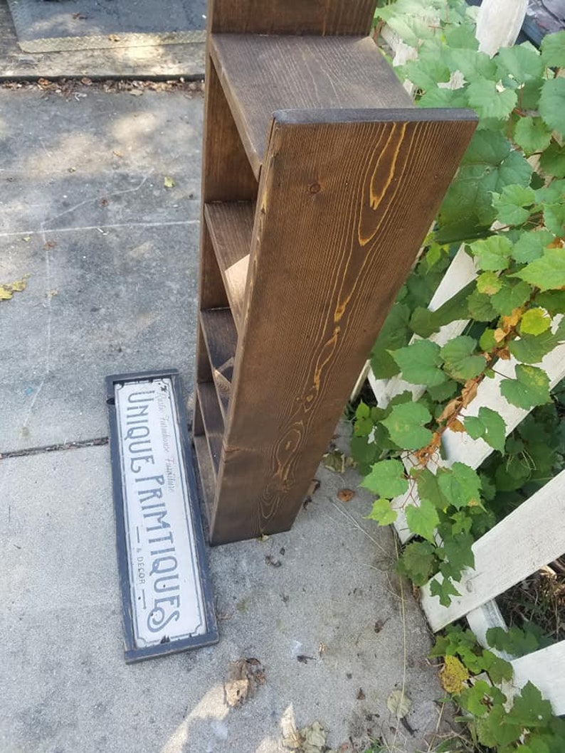 May include: A dark brown wooden shelf unit with four shelves. The shelf unit is standing on a gray concrete surface. A sign that reads "Unique Primitives - Home Decor" is lying on the ground in front of the shelf unit.