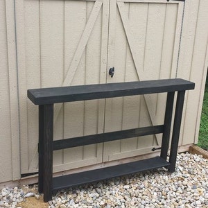 May include: A black wooden console table with two shelves. The table is standing in front of a shed with double doors.
