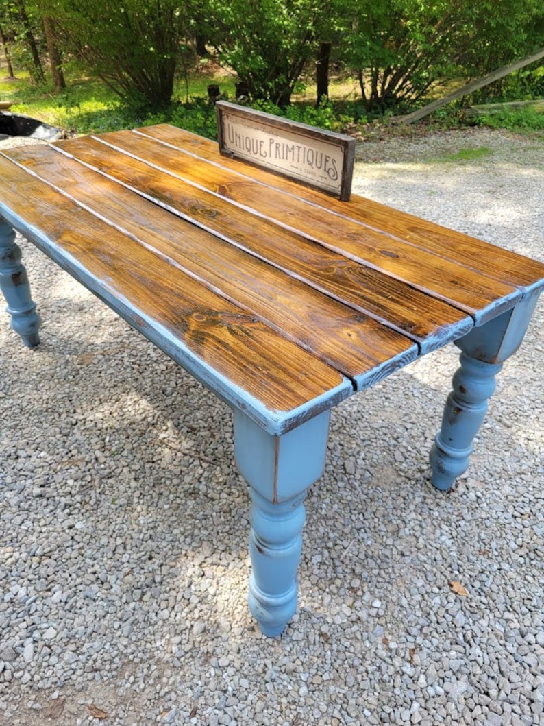 May include: A rustic wooden table with a stained brown tabletop and distressed blue legs. The table features a sign that reads "UNIQUE PRIMTIQUES". The table is made of wood and is suitable for indoor or outdoor use.