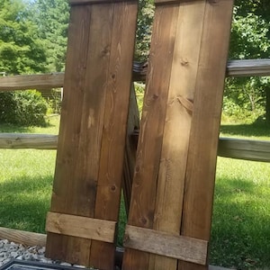 May include: Two wooden shutters with a rustic brown finish. The shutters are made of multiple planks of wood and have a simple design. The shutters are leaning against a wooden fence.