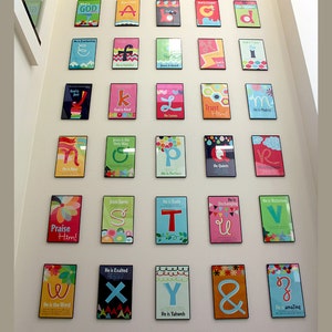 K&ouml;nnte beinhalten: Eine Wandanzeige mit bunten gerahmten Postern mit Buchstaben des Alphabets und religi&ouml;sen S&auml;tzen. Jedes Poster hat ein anderes Farbschema und Design, mit einem Buchstaben und einem Satz, der sich auf Gott oder Jesus bezieht. Die S&auml;tze beinhalten "Gott ist gerecht", "Er ist ewig", "Jesus ist gut", "Er ist nah", "Gott ist g&uuml;tig", "Er liebt", "Vertraue ihm", "Er ist m&auml;chtig", "Jesus ist der einzige Weg", "Er ist perfekt", "Er beruhigt", "Er ist siegreich", "Lobt ihn", "Jesus rettet", "Er ist das Wort", "Er ist erh&ouml;ht", "Er ist Jahwe", "Er ist erstaunlich".