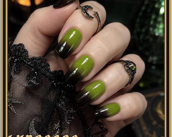 Puede incluir: Uñas con un diseño degradado que transita desde una base negra profunda hasta una punta verde oliva vibrante. Los dedos están adornados con dos anillos de metal, uno con forma de media luna y otro con una piedra redonda.