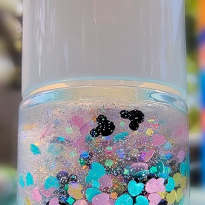K&ouml;nnte beinhalten: Eine klare zylindrische Flasche gef&uuml;llt mit einer Fl&uuml;ssigkeit, die mehrfarbige herzf&ouml;rmige Pailletten in Pink, Blau und Gelb sowie feinen silbernen Glitzer und zwei kleine schwarze Silhouetten in Form von Mausohren enth&auml;lt.