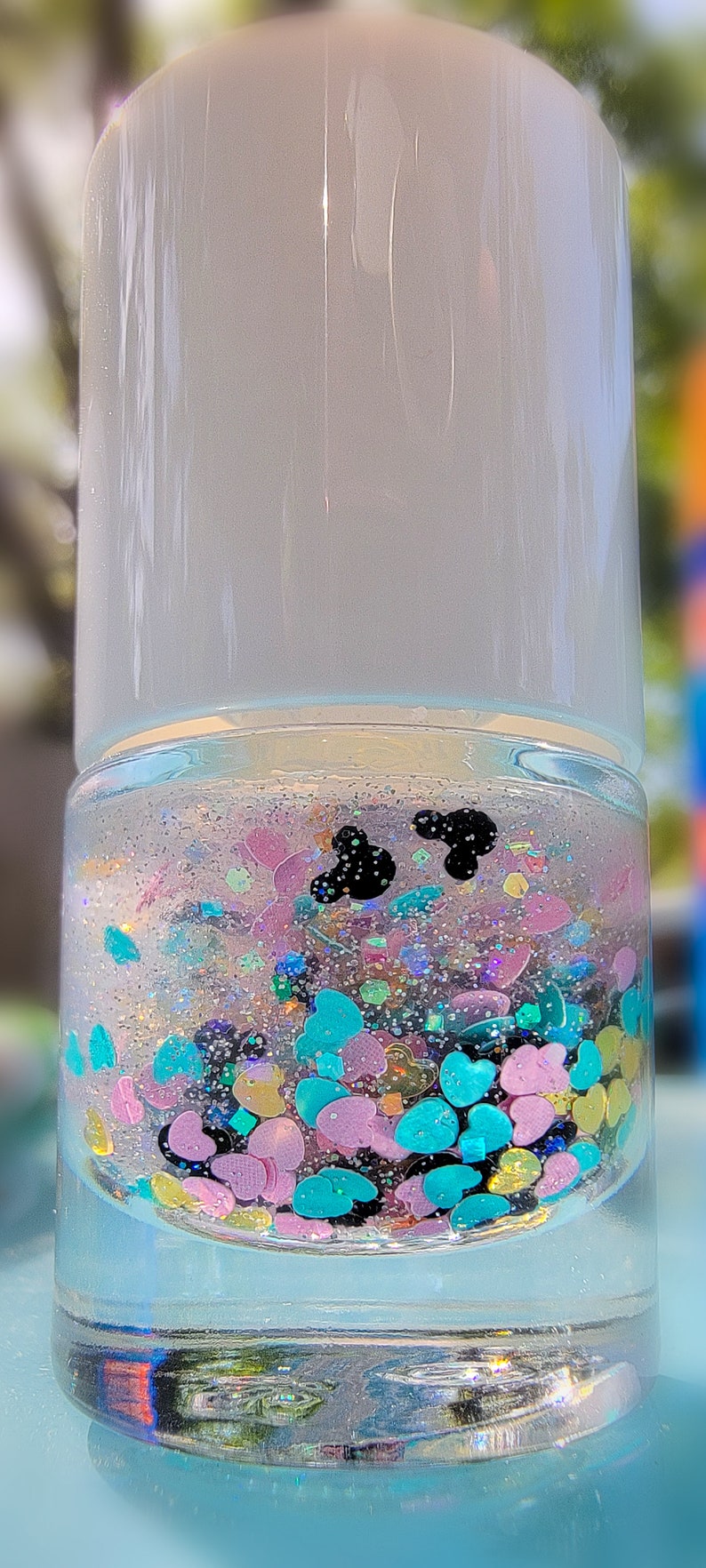 K&ouml;nnte beinhalten: Eine klare zylindrische Flasche gef&uuml;llt mit einer Fl&uuml;ssigkeit, die mehrfarbige herzf&ouml;rmige Pailletten in Pink, Blau und Gelb sowie feinen silbernen Glitzer und zwei kleine schwarze Silhouetten in Form von Mausohren enth&auml;lt.